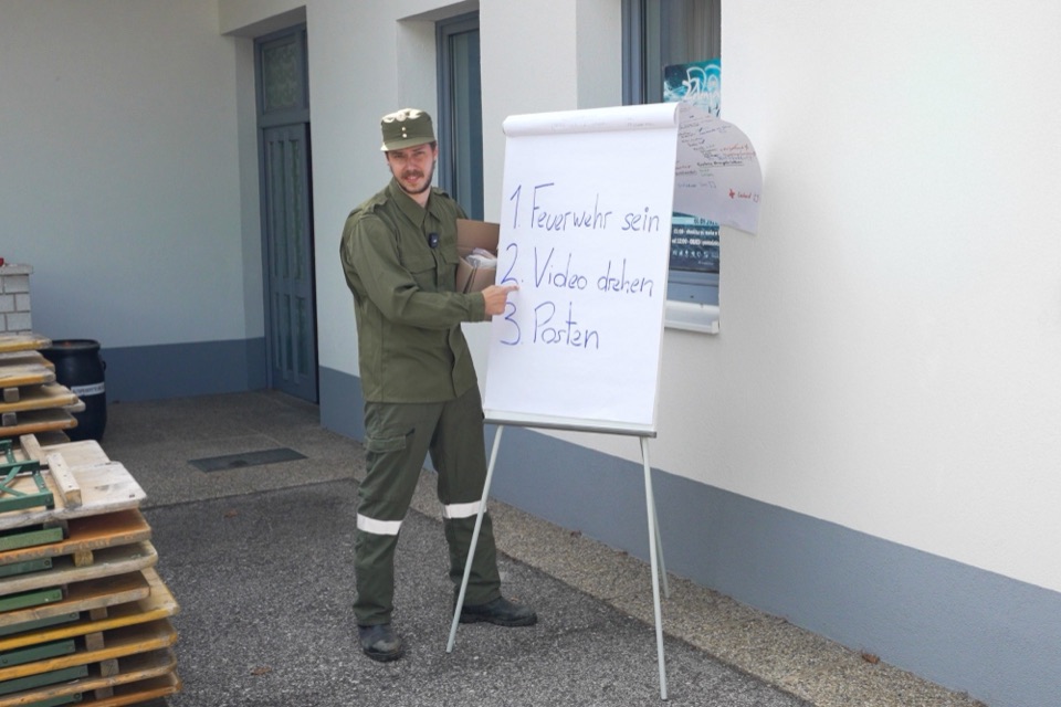 Feuerwehrmann in Uniform zeigt auf ein Flipchart: 1. Feuerwehr sein, 2. Video drehen, 3. Posten